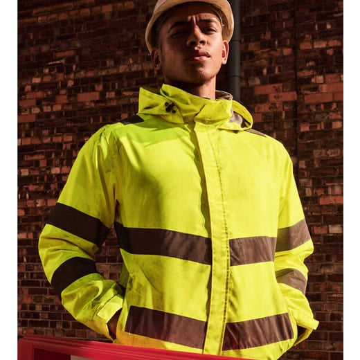 A man wearing a high-visibility neon yellow safety jacket with reflective brown stripes and a beige hard hat, standing in front of a brick wall.