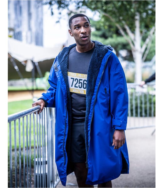 A man wearing a bright blue hooded raincoat with a dark fleece-lined interior, standing outdoors near a metal railing. He is wearing a black t-shirt and a race bib with the number 7256.