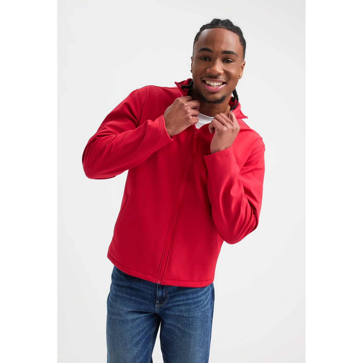 A man wearing a bright red zip-up hoodie and blue jeans, smiling and adjusting the collar of the hoodie against a plain light gray studio background.