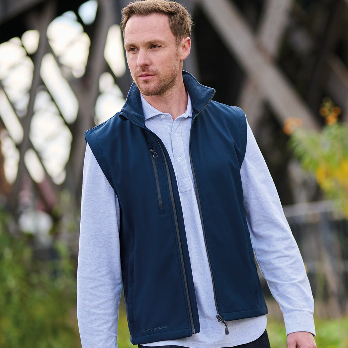 A man wearing a navy blue zip-up vest over a light blue long-sleeve polo shirt, standing outdoors in front of a blurred bridge structure.