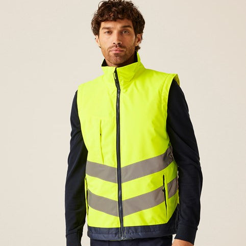 A man wearing a high-visibility neon yellow safety vest with reflective silver stripes over a dark navy long-sleeve shirt and navy trousers, photographed against a plain light beige studio background.